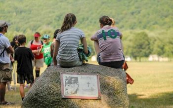 Two girls sit on a large rock that has a plaque on it with a photo of a priest.