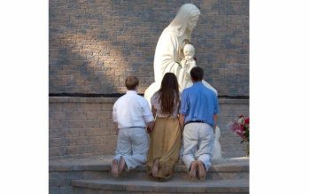 Three young people kneel in prayer before a statue of Mary holding the infant Jesus at an outdoor shrine.